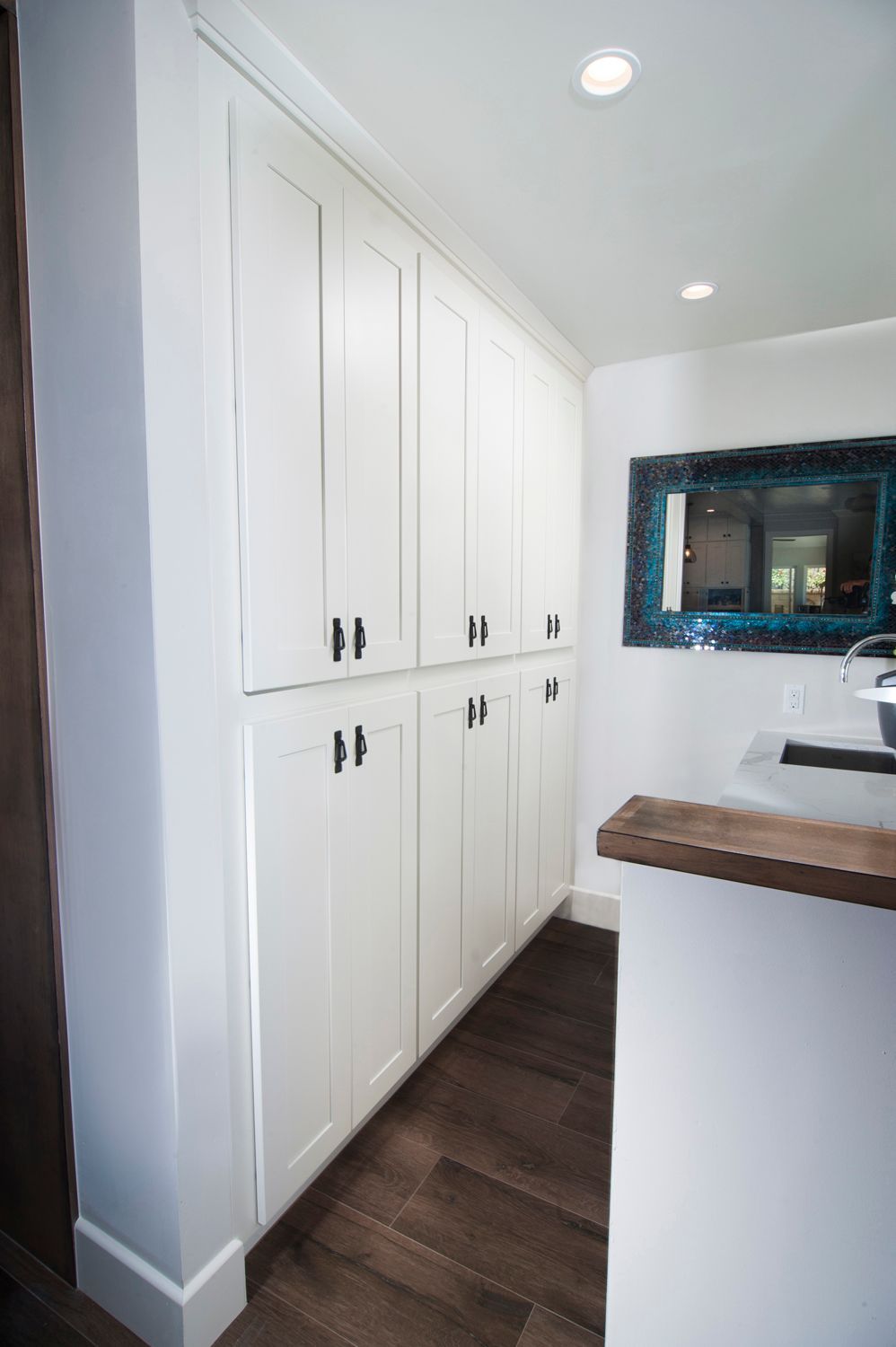 White cabinets with black handles in a kitchen. Dark wood floor, white walls, a mirror.