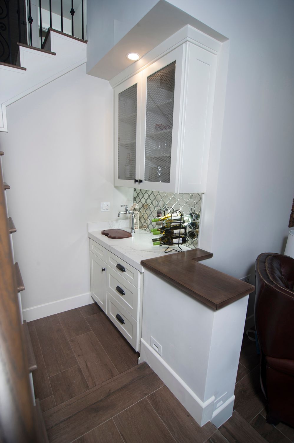 White built-in bar area under stairs, including cabinets, countertop, backsplash, and dark wood accents.