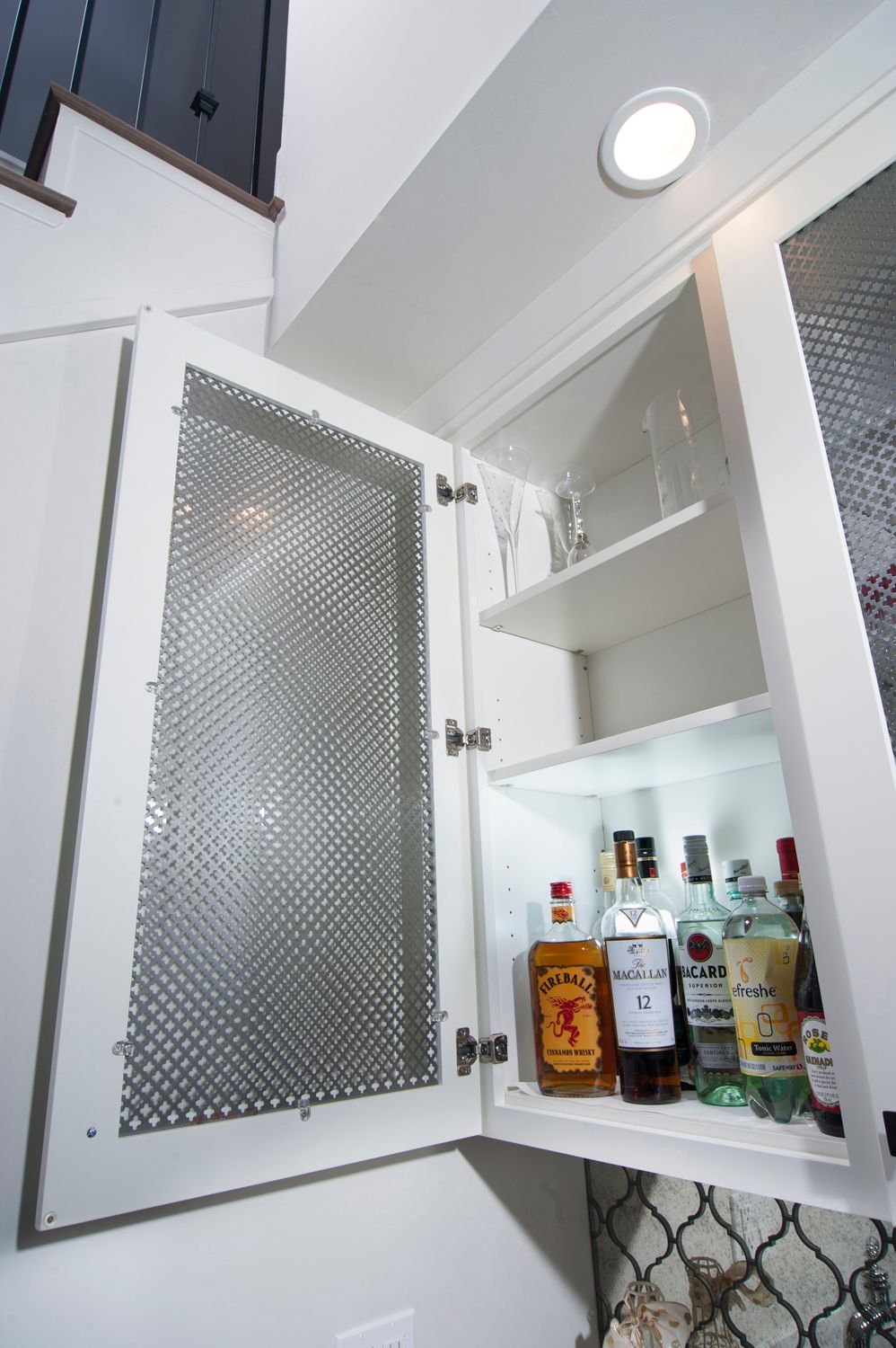 White cabinet with open door showing liquor bottles, glassware, and a textured metal panel.
