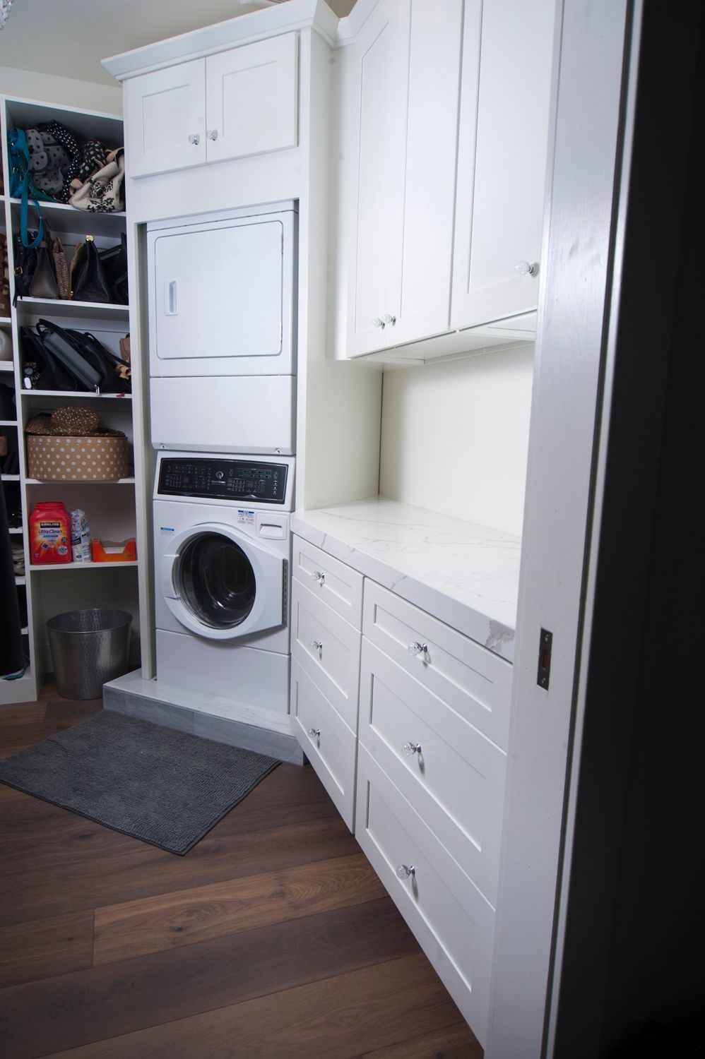 White laundry room with stacked washer/dryer, cabinets, drawers, and wood floor.