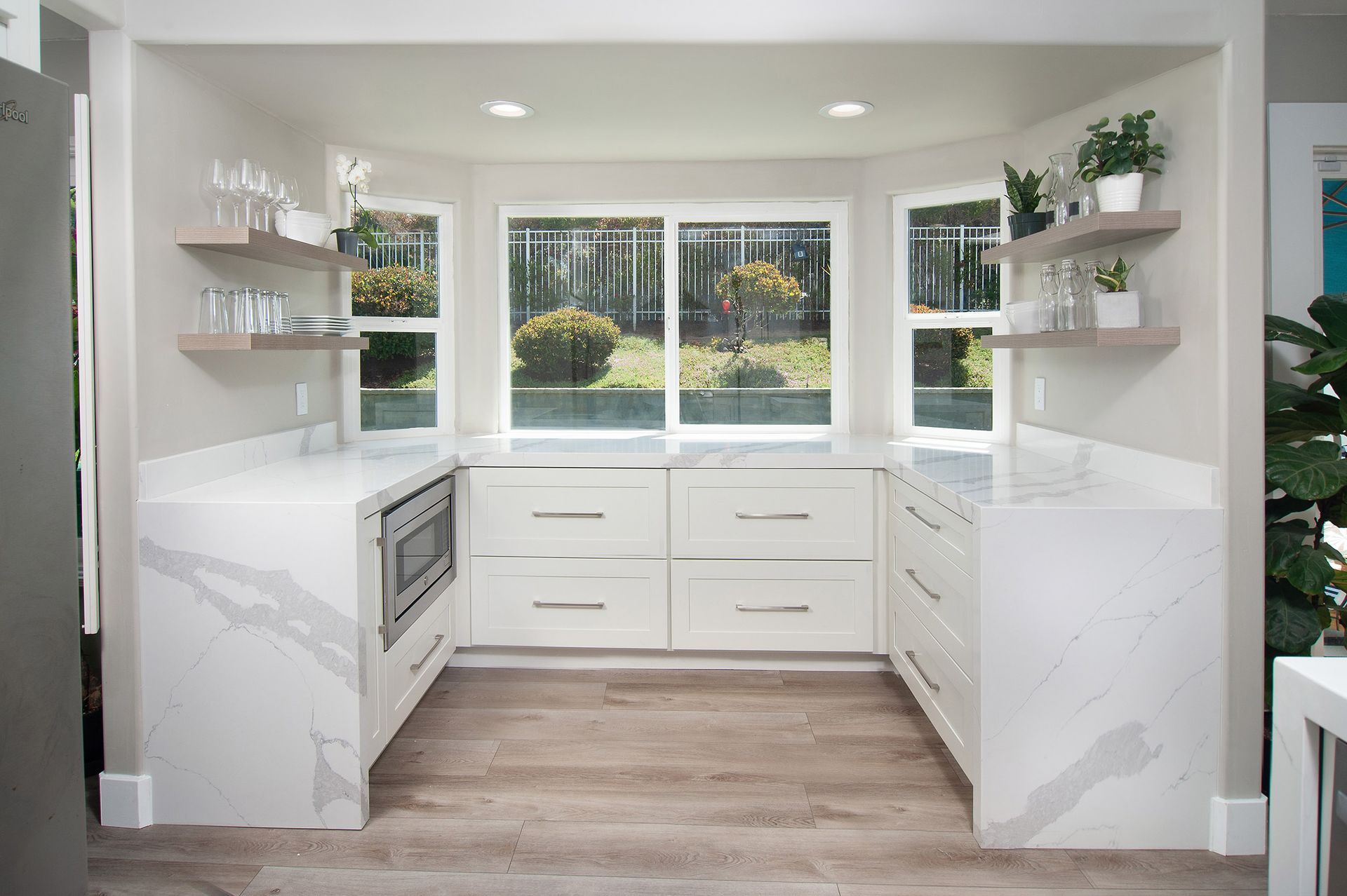 U-shaped white kitchen with quartz countertops, built-in microwave, and a large window overlooking greenery.