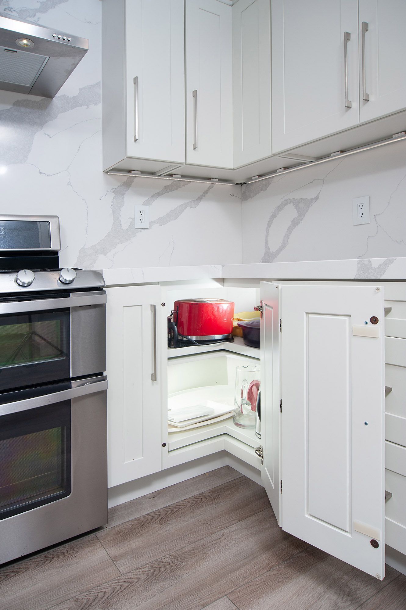 White kitchen corner cabinet open, revealing a red pot. Stainless steel oven and white countertops.