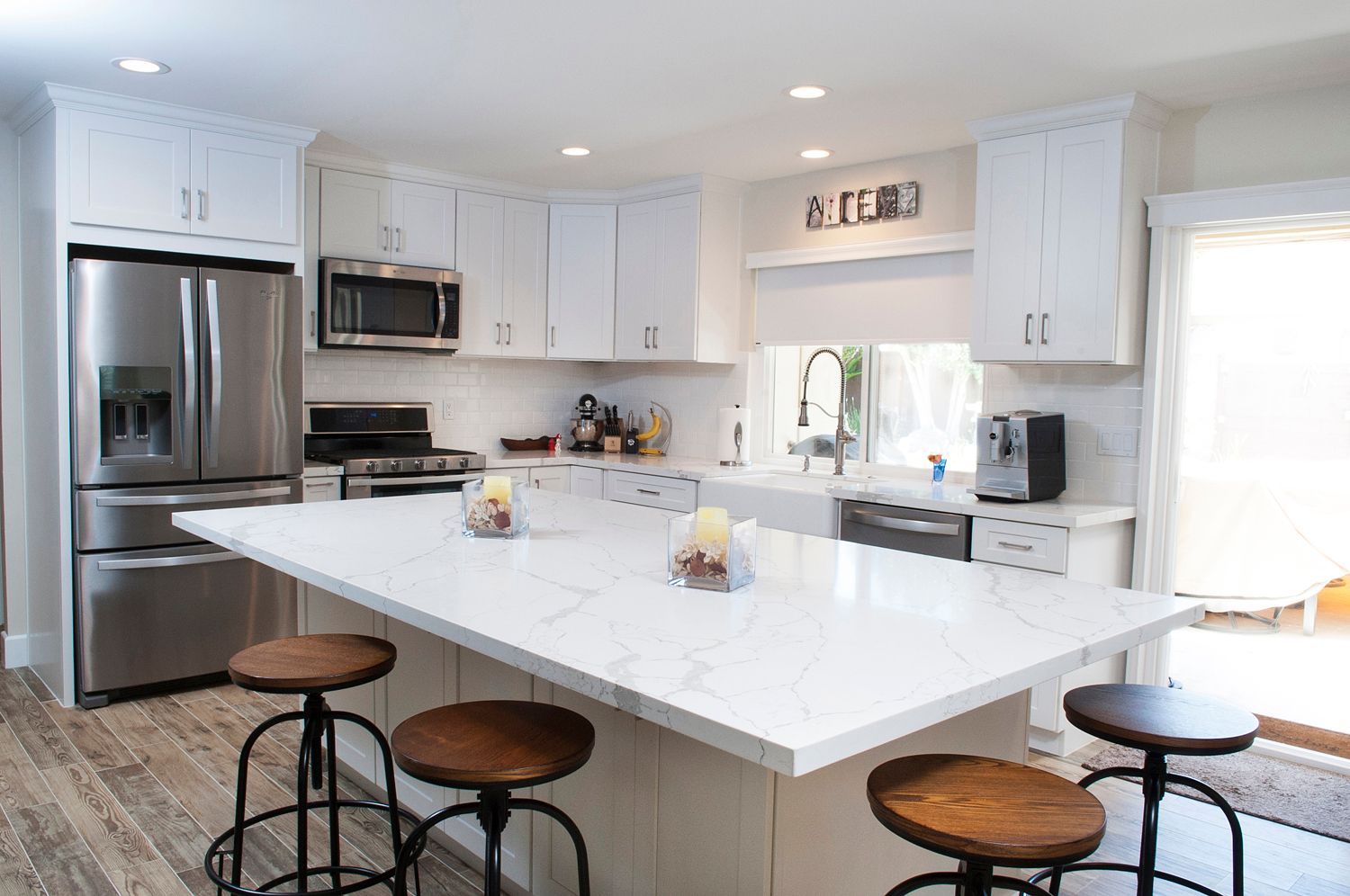 White kitchen with island, stainless steel appliances, wooden stools, and natural light.