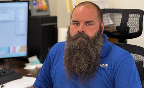 Man with a long beard in a blue shirt sitting at an office desk, looking at the camera.