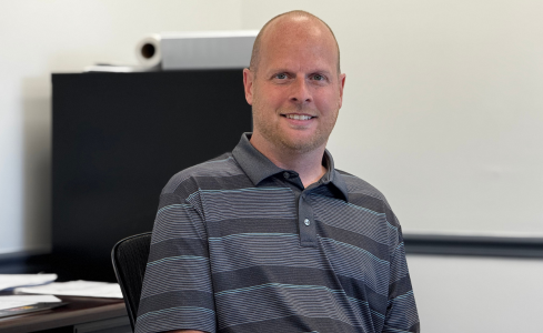 Man seated at a desk in an office, wearing a gray striped shirt and smiling at the camera