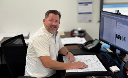 Man sitting at a desk reviewing a large plan on paper beside two computer monitors in an office
