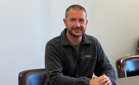 Man seated in a chair, smiling in a gray sweater against a light wall.