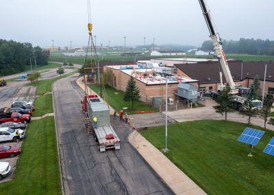 Crane lifts a white truck over a road beside a building and parking lot on an overcast day