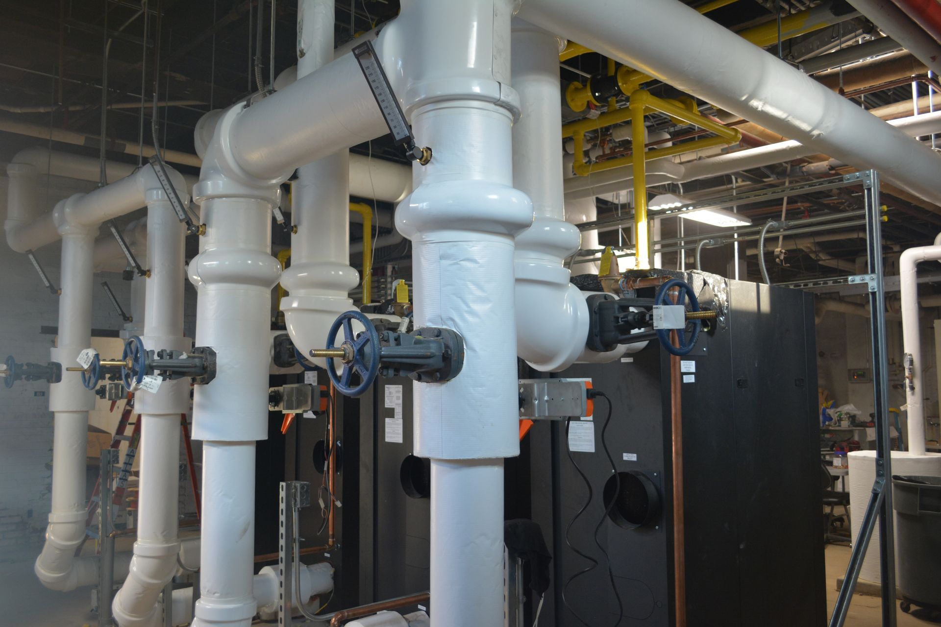 Industrial facility with large white pipes, valves, and black equipment in a mechanical room