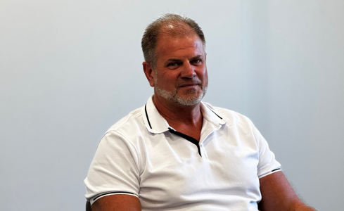 Man in a white polo shirt seated against a plain light-gray background, looking slightly off-camera.
