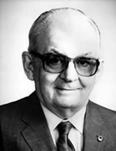 Portrait of a man in a suit and tie, wearing glasses, against a plain background