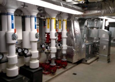 Industrial basement with white and red pipes, valves, and insulated ductwork along a mechanical room wall