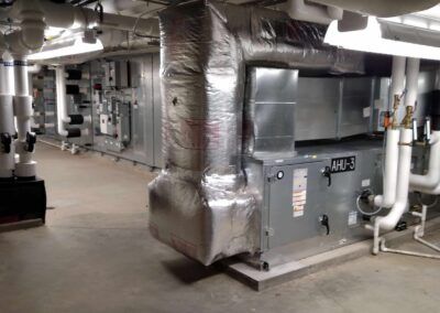 Industrial HVAC ductwork and equipment in a basement mechanical room with metal piping and insulation.