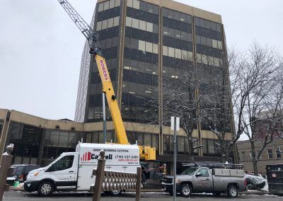 Crane lifting equipment beside a dark office building on a snowy street, with parked vehicles below.