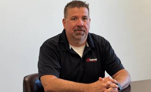 Man seated at a table in a black polo shirt, facing the camera in a plain office setting
