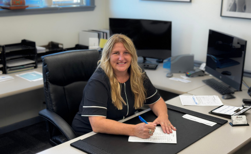 Woman smiling at an office desk, writing on paper with computers and files nearby.