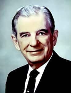 Formal portrait of a smiling man in a dark suit and tie against a light blue background