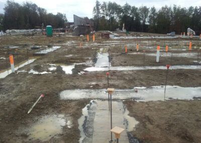 Construction site with muddy ground, white survey markings, orange cones, and a small concrete footing area