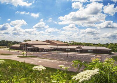 Large brown building with a parking lot under a bright cloudy sky, viewed from a grassy hill