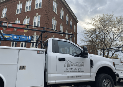 White utility truck with ladder parked beside a brick building on a city street under cloudy skies