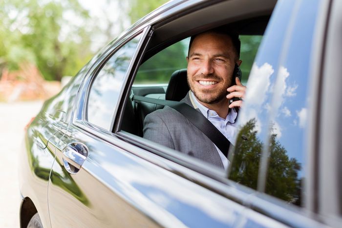Smiling man in a gray suit talking on a phone inside a black car, seen through the open window