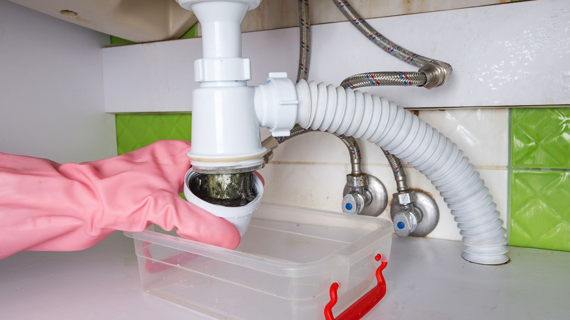 Person wearing pink glove removing a sink trap over a container, under a sink.
