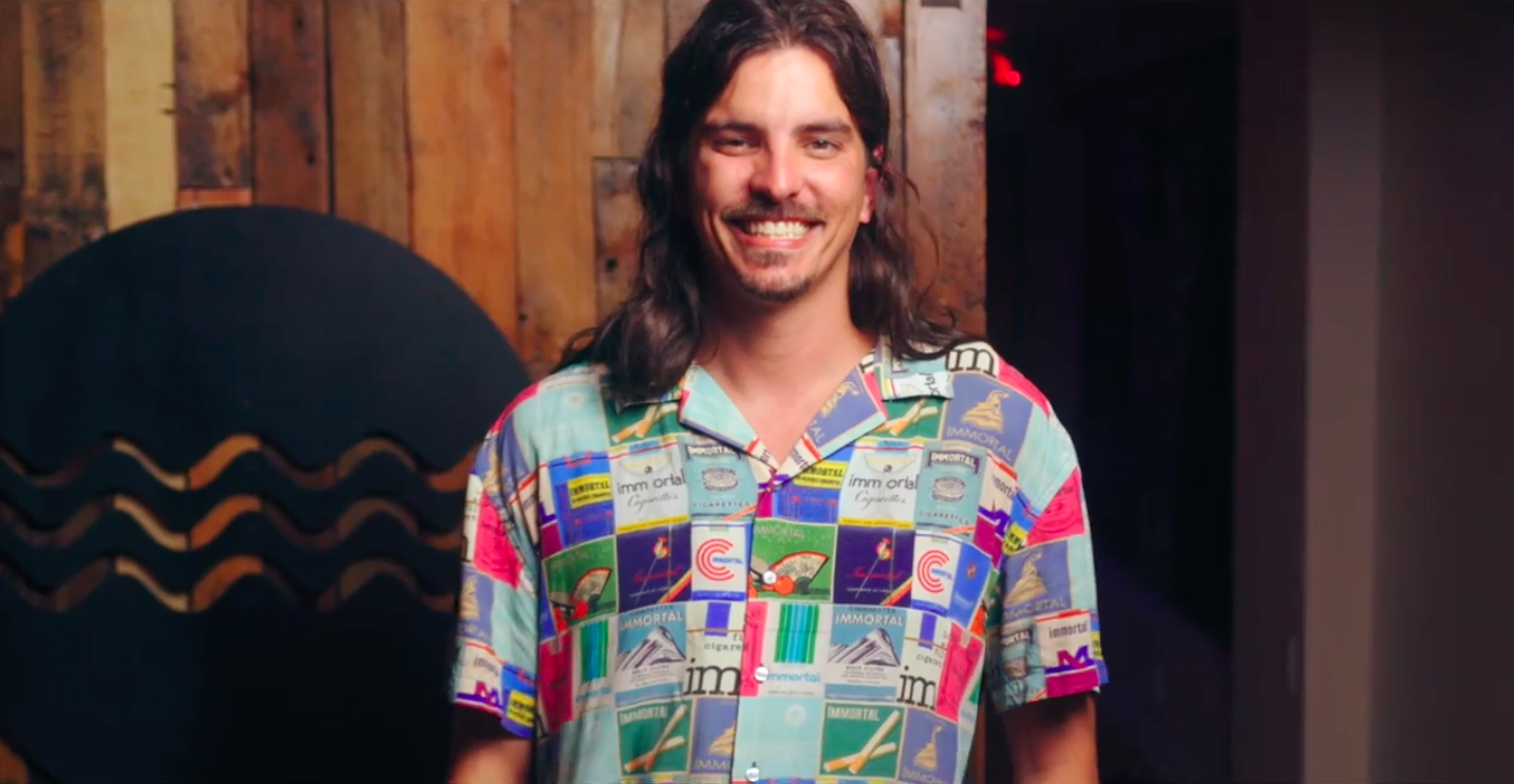 A man with long hair is wearing a colorful shirt and smiling.