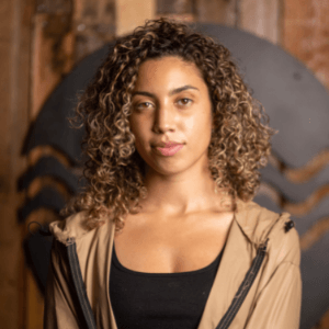 A woman with curly hair is wearing a brown jacket and a black top.