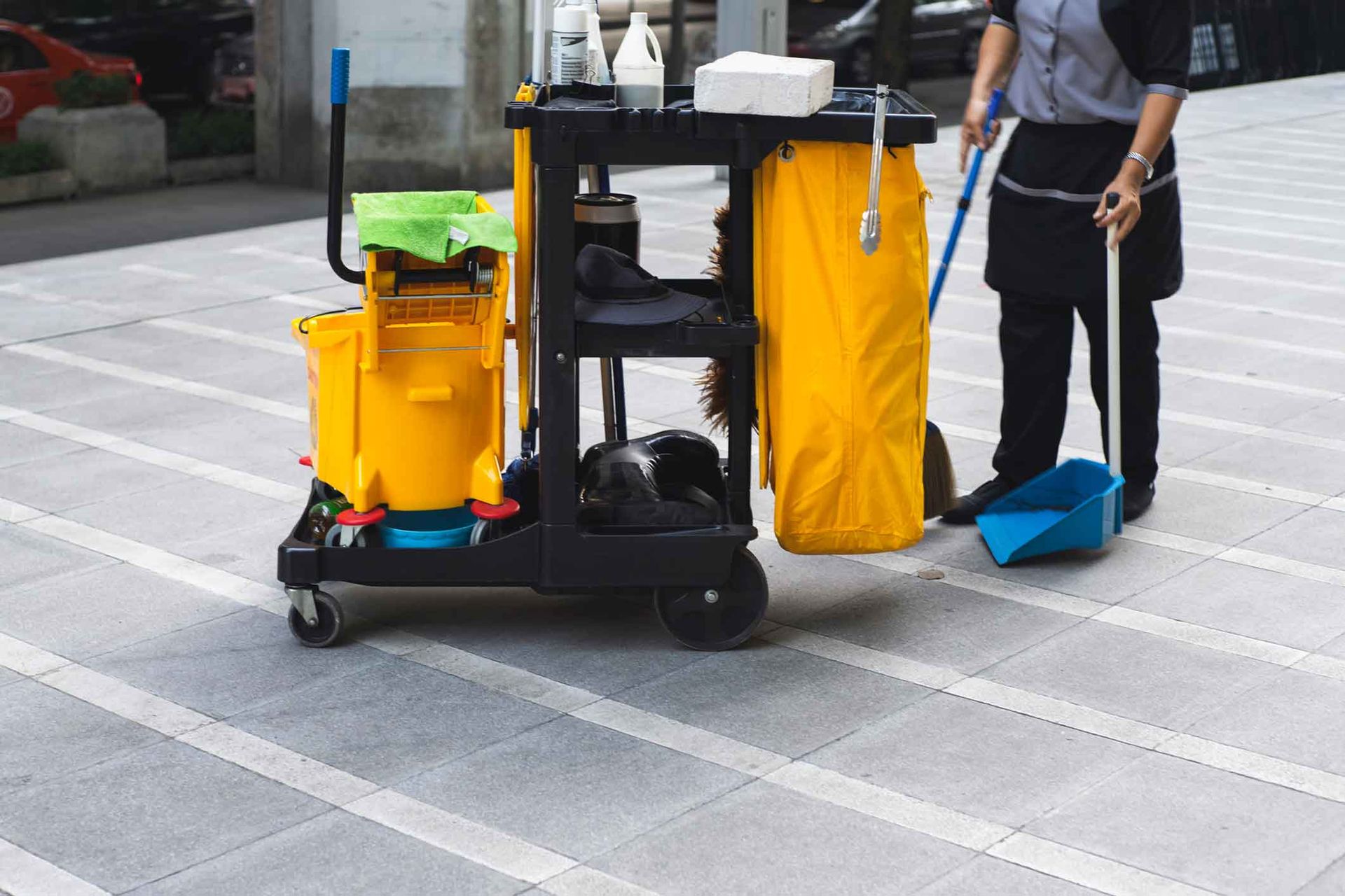 A woman is standing next to a cleaning cart on a sidewalk.