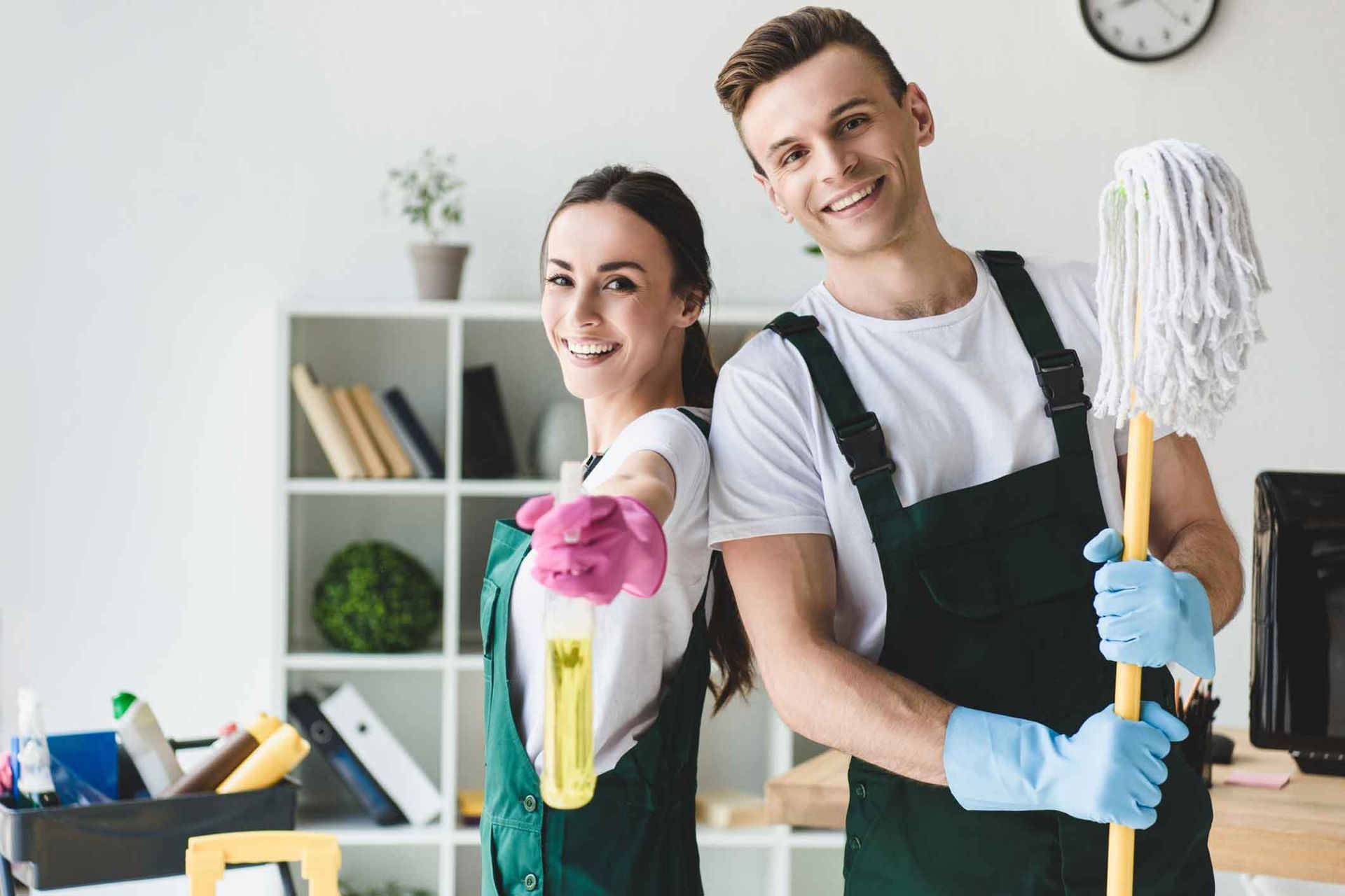 A man and a woman are standing next to each other in an office holding cleaning supplies.