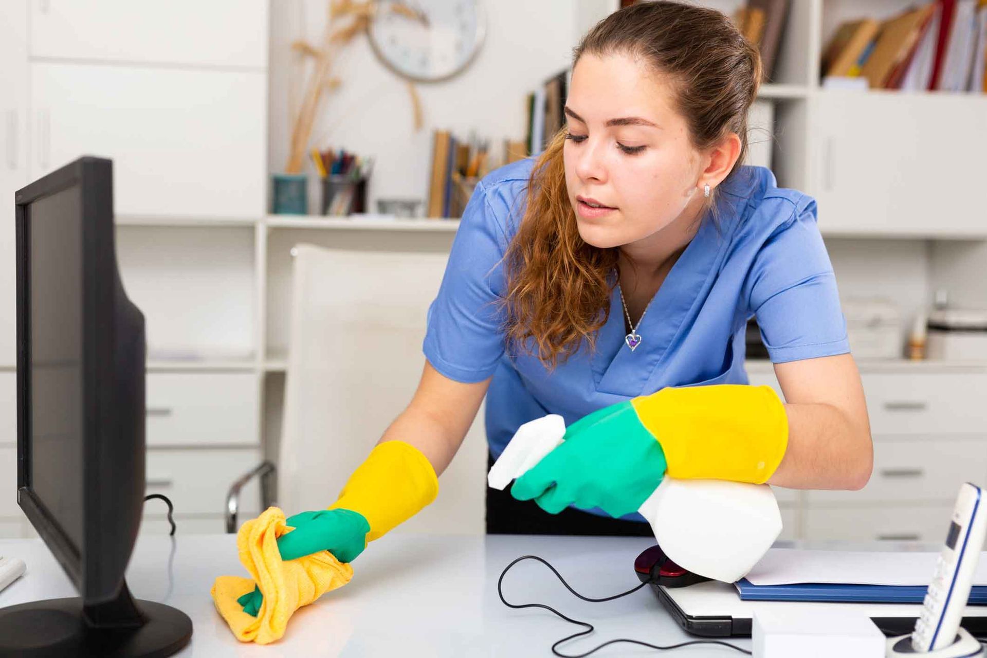 A woman is cleaning a desk with a cloth and spray bottle.