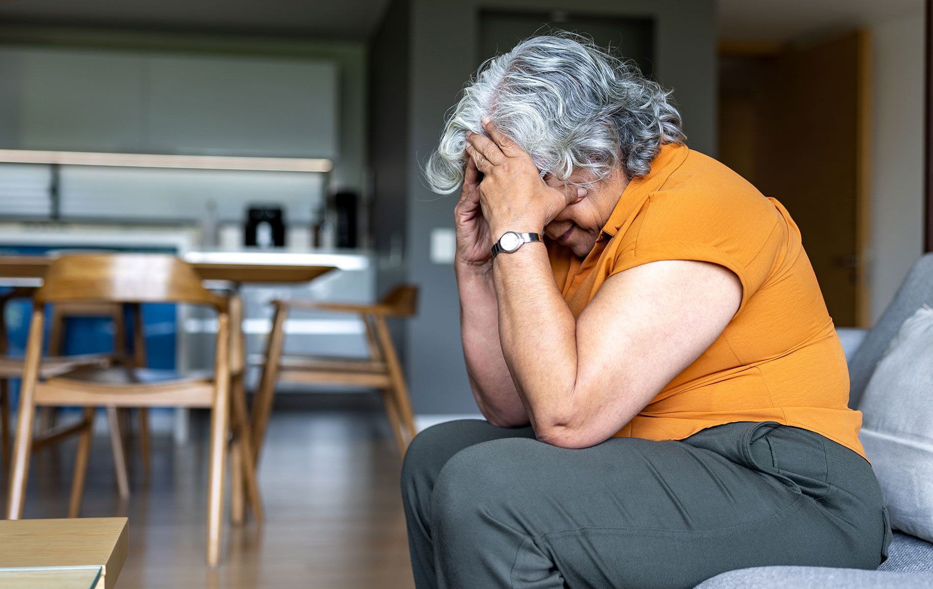 An elderly woman is sitting on a couch with her head in her hands.
