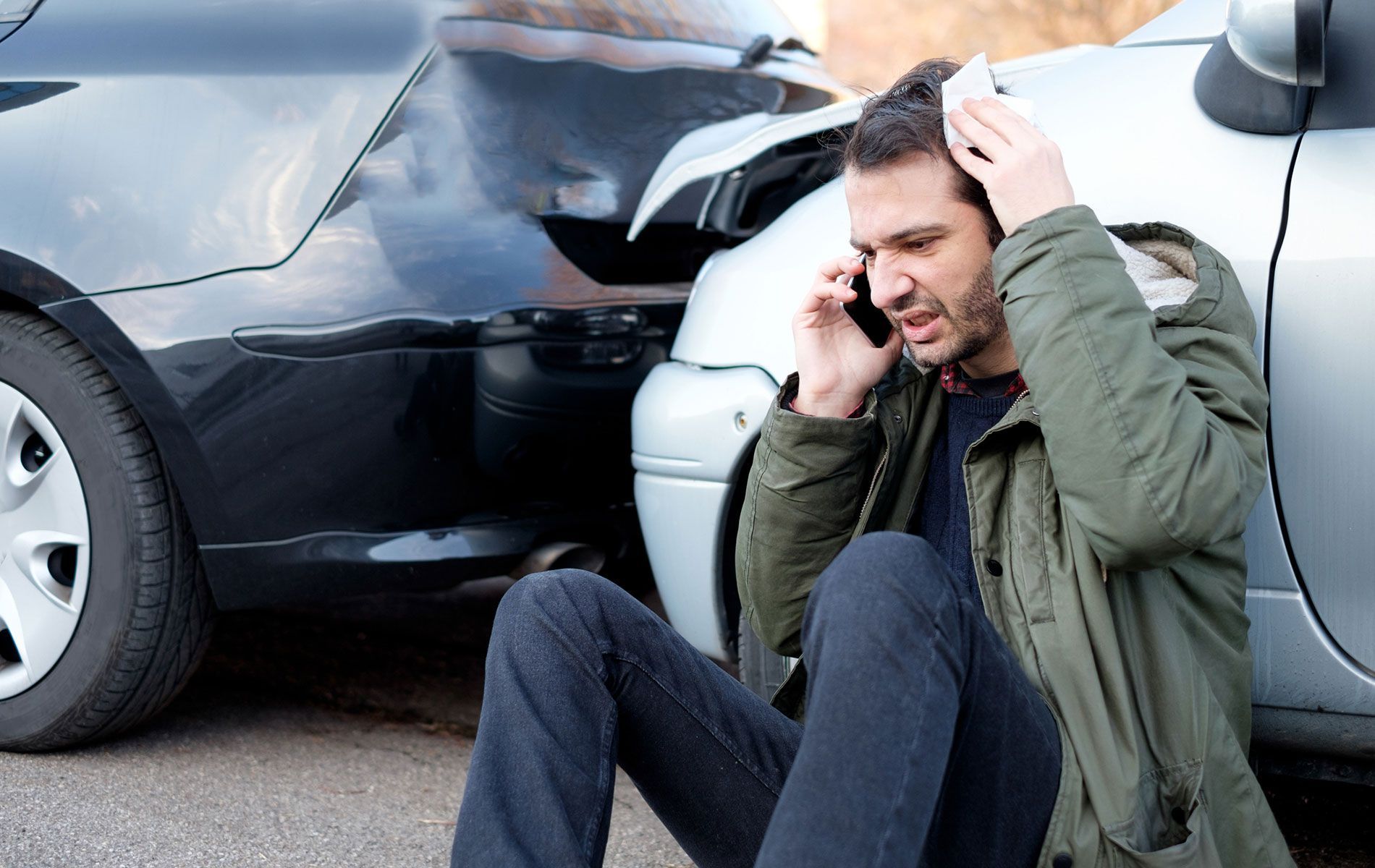 A man is sitting on the ground talking on a cell phone in front of a car accident.