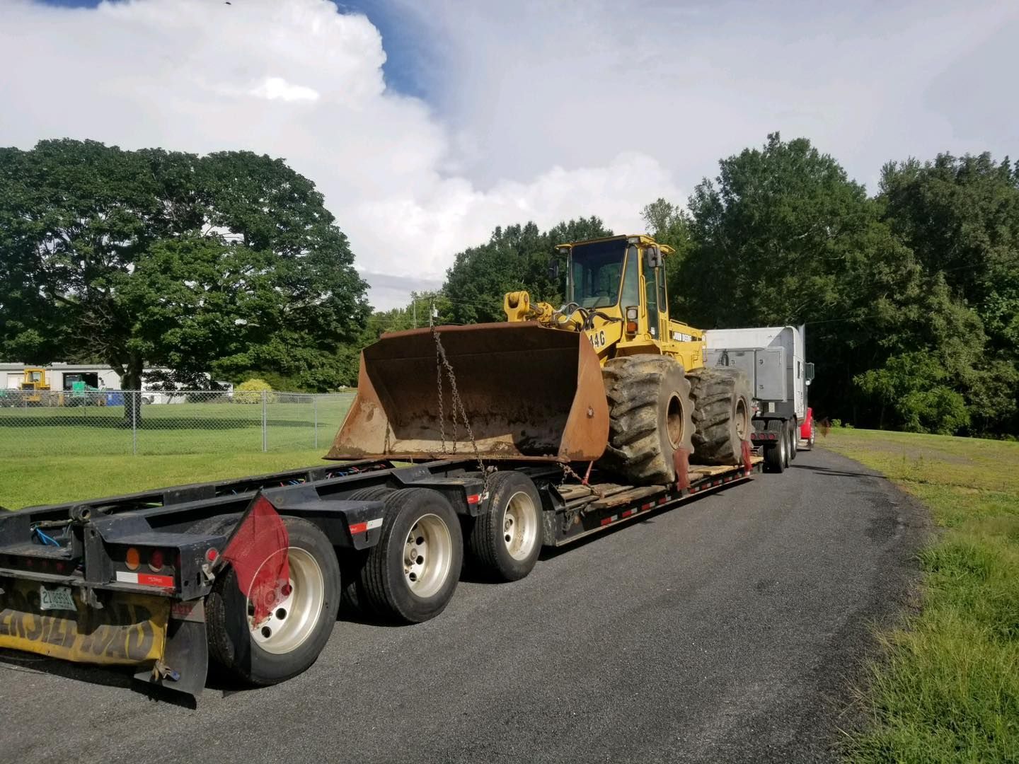 Yellow earthmover on a black flatbed trailer on a gravel road, trees in the background.