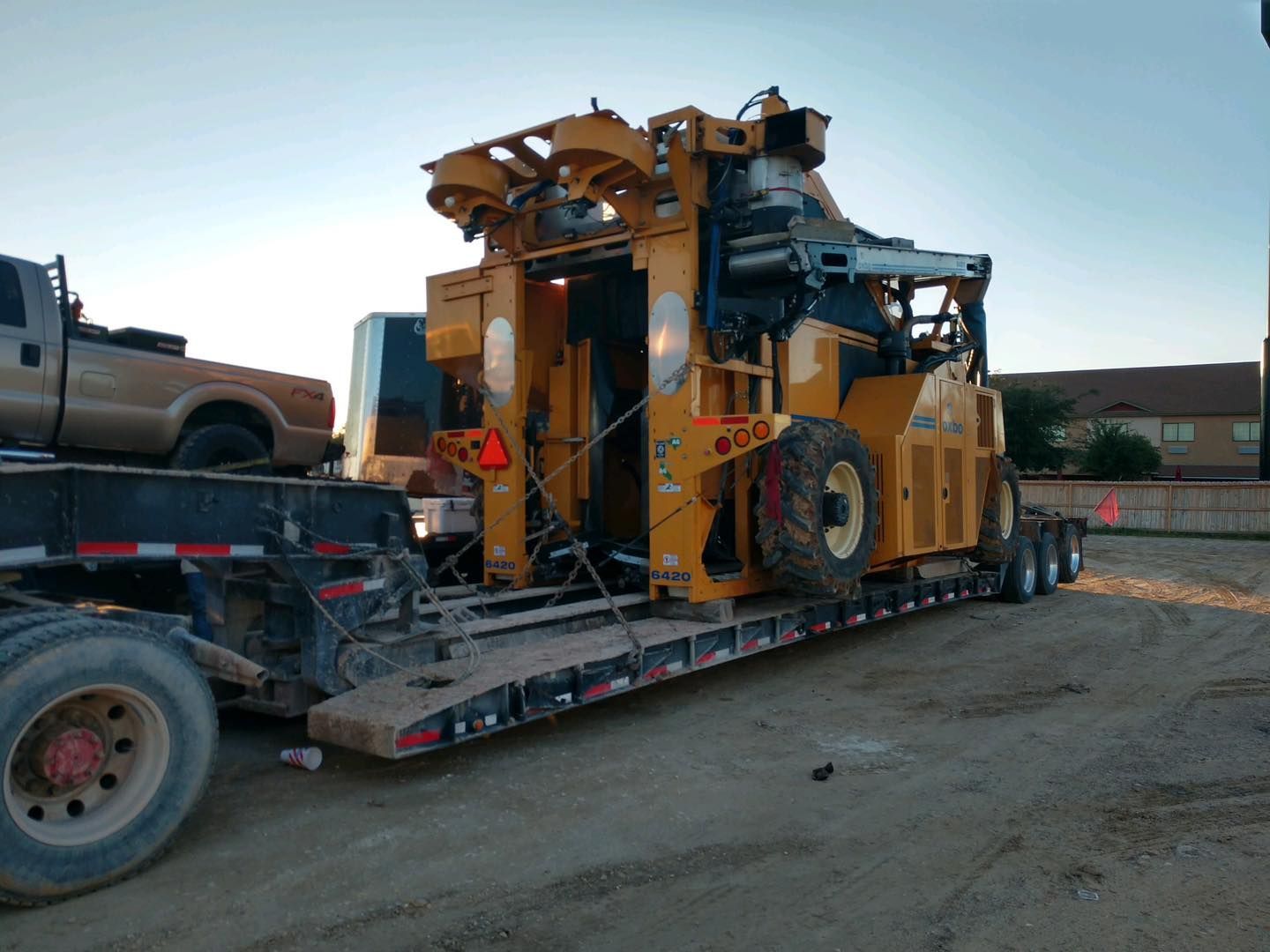 Yellow industrial machine on a flatbed trailer, backed by a pickup truck. Daylight setting.