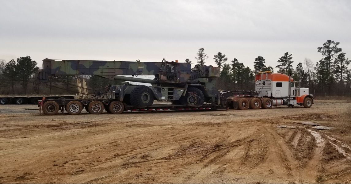 Crane loaded on a flatbed trailer, pulled by a semi-truck on a dirt road.