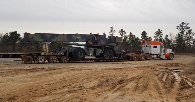 Crane loaded on a flatbed trailer, pulled by a semi-truck on a dirt road.