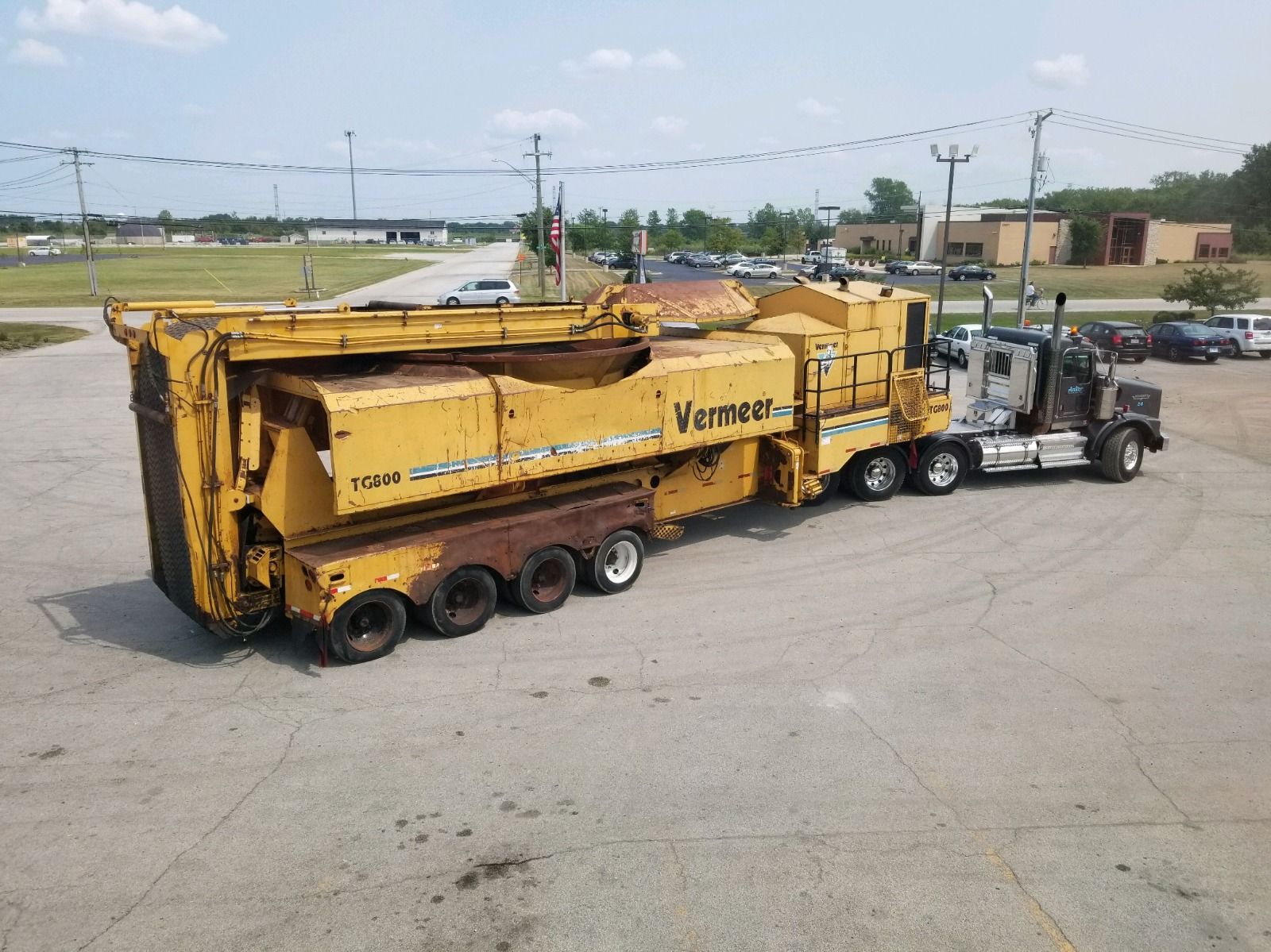 Yellow Vermeer wood grinder and semi-truck on pavement; sunny day.