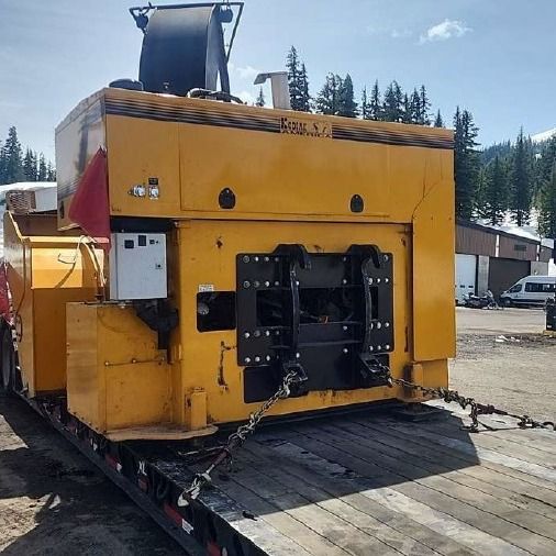 Yellow snow-making machine on a flatbed trailer, chained down, outdoors.