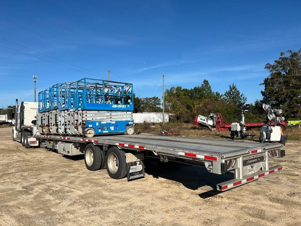 Flatbed truck carrying stacked blue scissor lifts under a clear, bright sky.