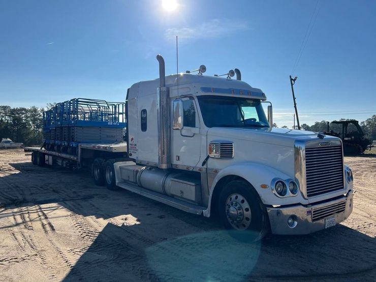 White semi-truck with flatbed trailer carrying blue metal frames on a dirt lot under a sunny sky.