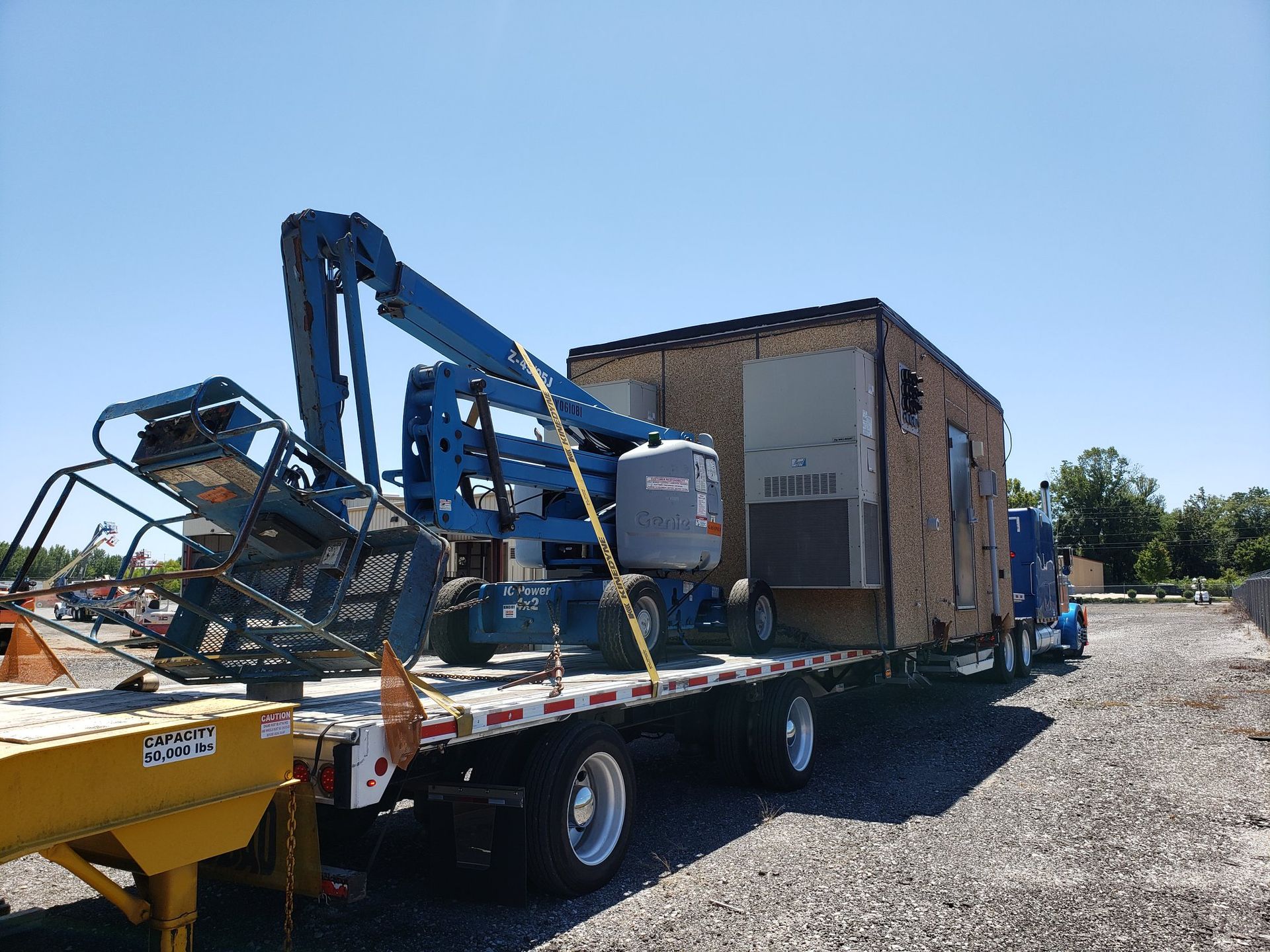 Flatbed truck transporting a large brown structure with a blue lift on a sunny day.