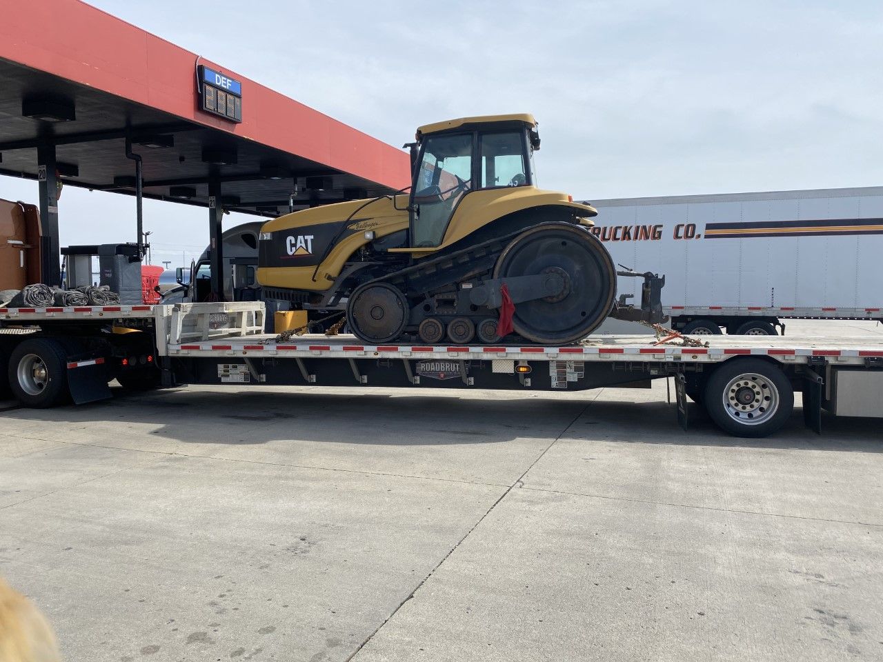 Yellow Caterpillar tractor on a flatbed trailer at a gas station under a red canopy.