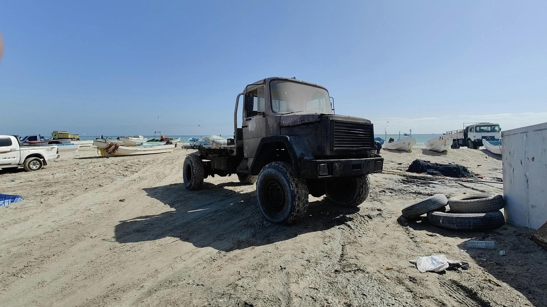 Black truck on a sandy beach under a blue sky, surrounded by debris.