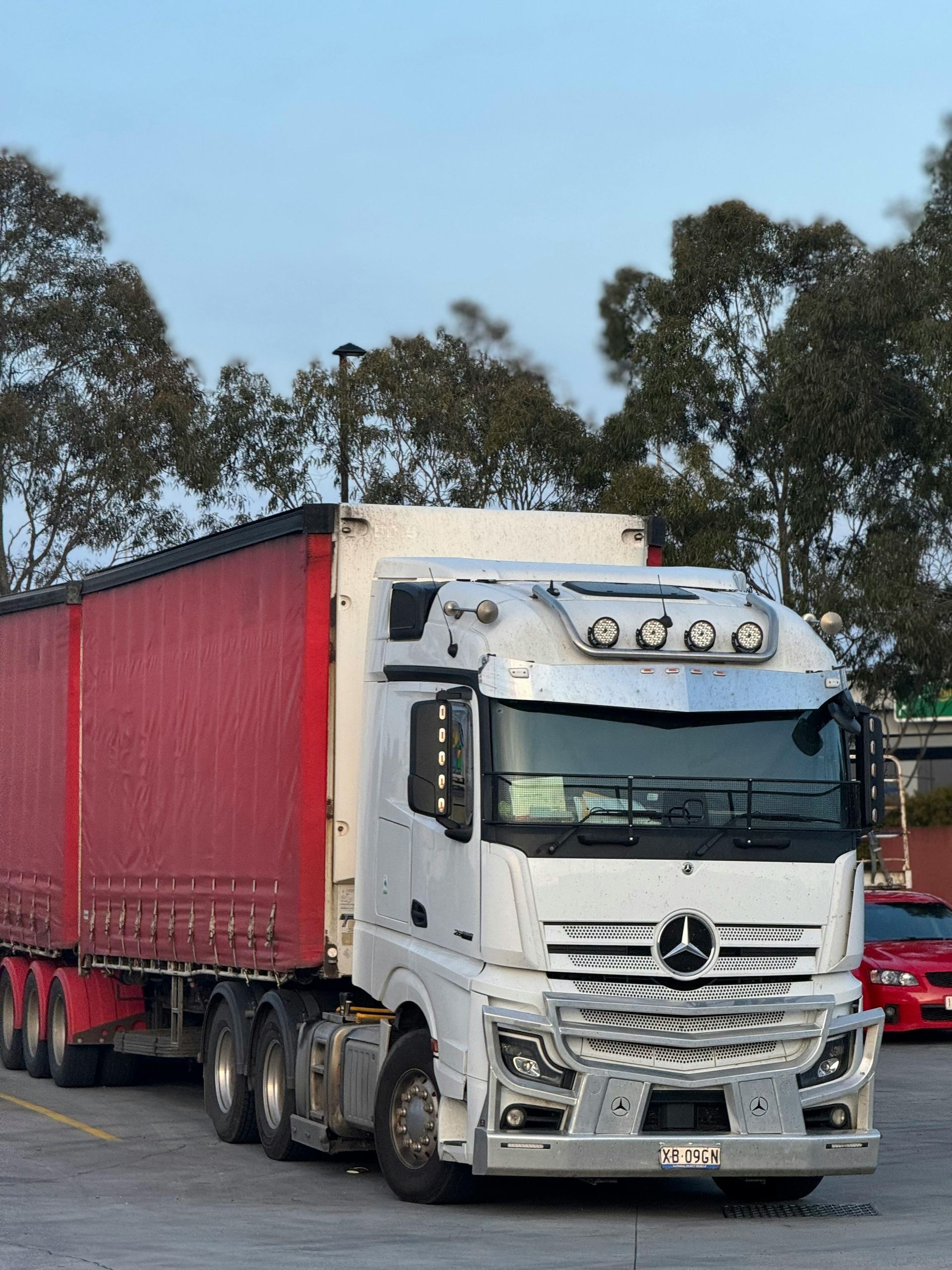 White Mercedes semi-truck with a red trailer parked outdoors under a cloudy sky.