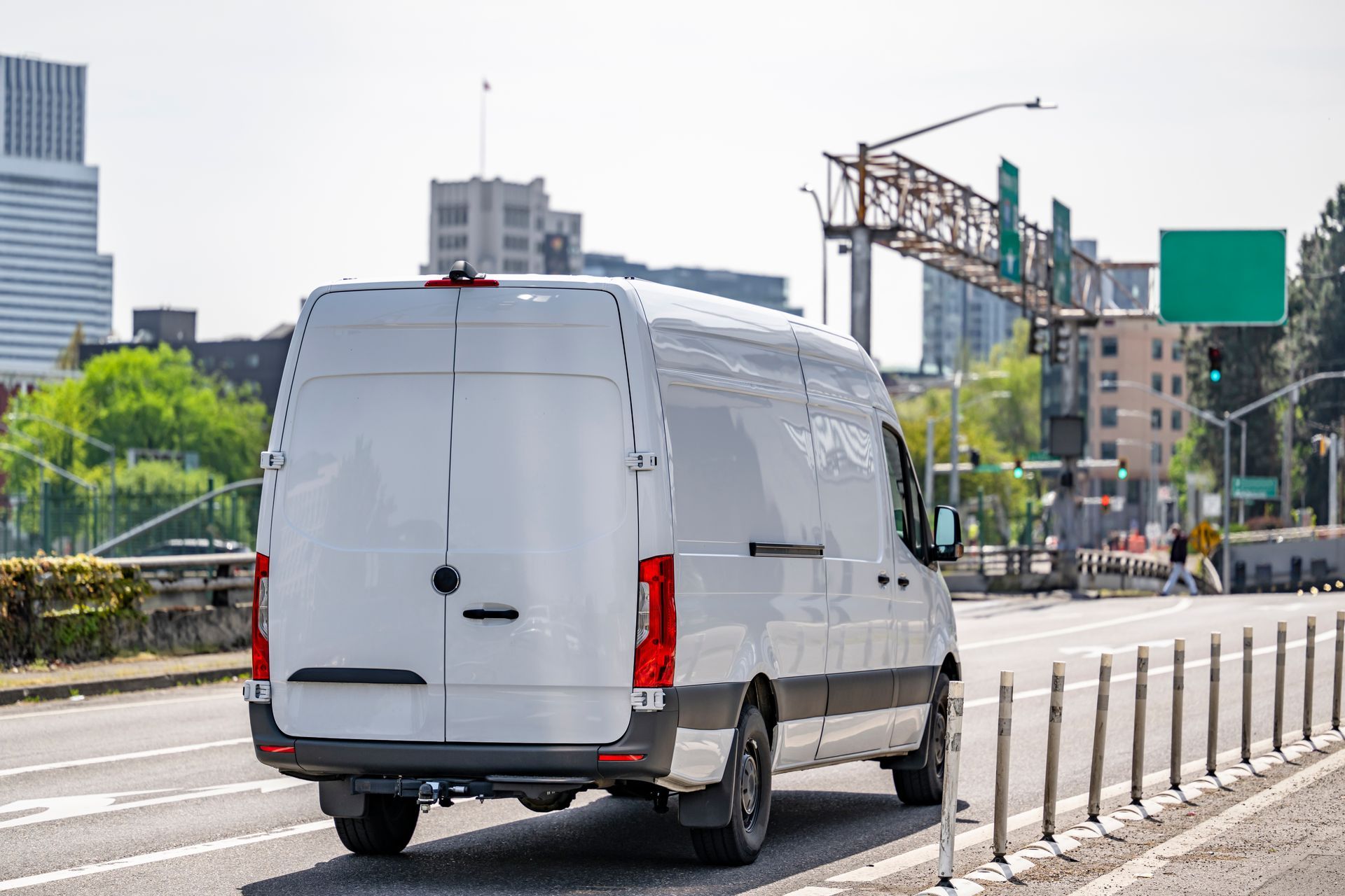 White cargo van driving on a city road. Buildings and a traffic signal are visible.