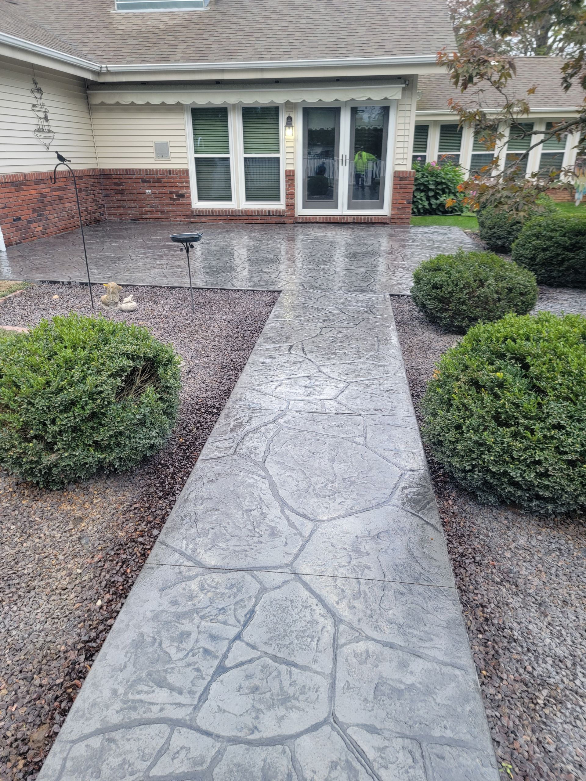 a grey stamped concrete walkway leading to a house surrounded by bushes and gravel .