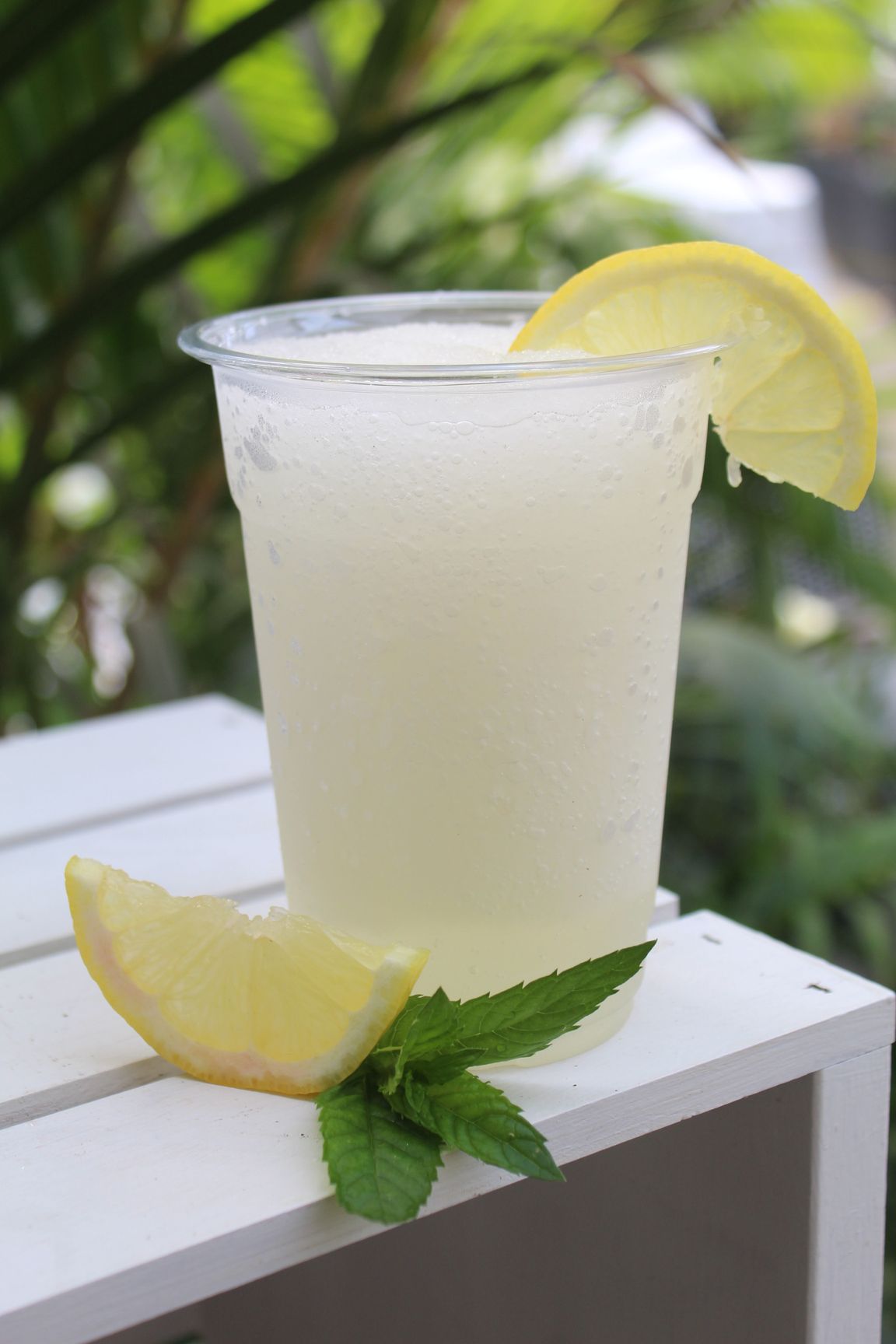 Lemonade in a clear plastic cup with lemon slice garnish and mint on a white wooden surface.
