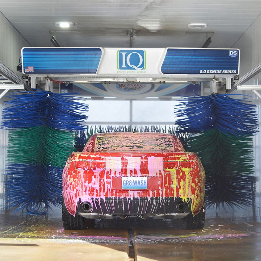 Red car in a car wash, covered in colorful soap, with blue and green brushes.
