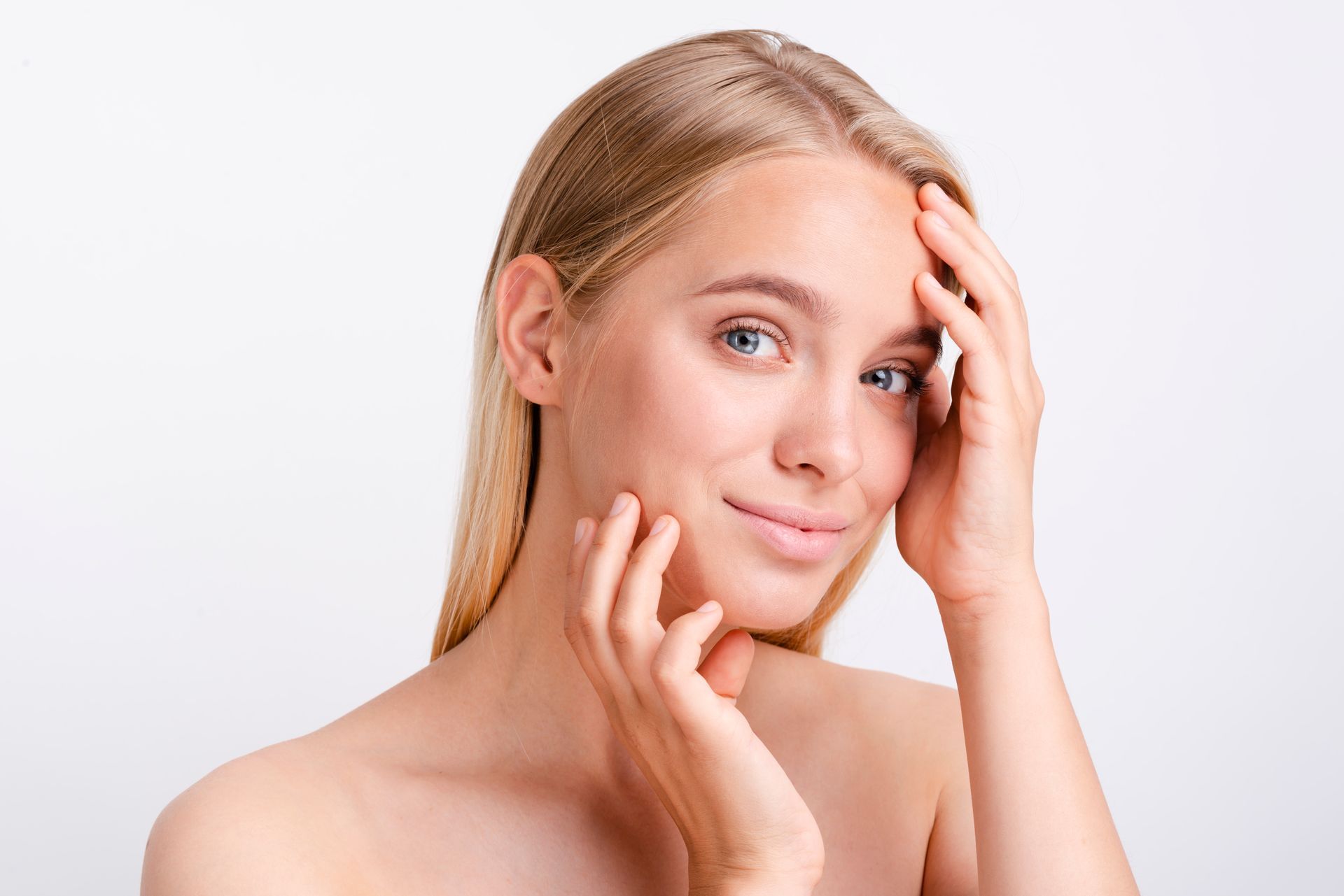 Blonde woman with hands touching face, smiling, against white background.